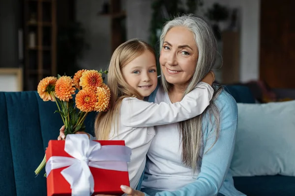Cheerful european small girl hugging old woman with gift box and bouquet of flowers in living room interior. Grandma and granddaughter relationship, congratulations on holiday and birthday celebration