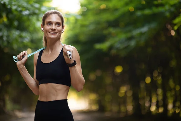 Training equipment concept. Happy athletic blonde sporty woman in black sportswear exercising at public park, using jumping rope, looking at copy space and smiling, panorama, closeup shot