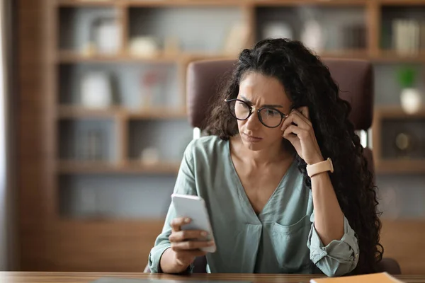 Concentrated millennial woman in casual outfit with eyeglasses sitting at workdesk at office, using brand new cell phone, reading post on Internet, using business mobile app, touching head, copy space