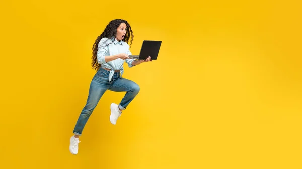 Shocked Arabic Woman Holding Laptop Running In Mid Air And Using Computer Working Online Posing Over Yellow Studio Background. Panorama With Blank Space For Text