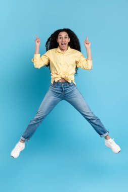 Joyful Arabic Female Jumping Pointing Fingers Up Posing In Mid Air Advertising Great Offer Over Blue Studio Background. Check This Out Concept. Vertical, Full Length Shot
