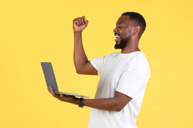 Cheerful Black Male Using Laptop Gesturing Yes Celebrating Success Browsing Internet Standing Over Yellow Studio Background. Side View Shot. Internet And Technology Concept