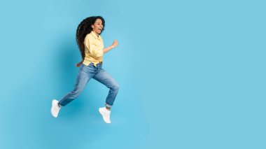 Cheerful Middle Eastern Woman Running Posing In Mid Air Jumping Over Blue Studio Background. Panorama With Blank Space For Text. Side View, Full Length Shot