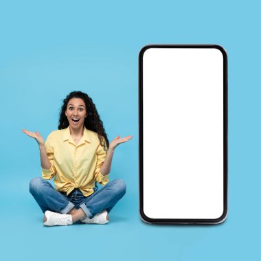 Excited young woman sitting near big cellphone with empty space for mobile app or website on screen over blue studio background, mockup. Great promo, online offer concept
