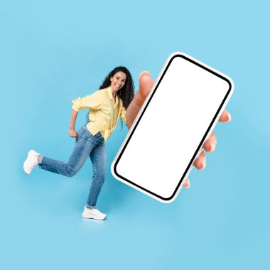 Cheerful Middle Eastern Lady Showing Huge Smartphone With Blank Screen Advertising Application Posing In Studio Over Blue Background. Technology And Gadgets Concept. Mockup