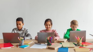 Three Multiracial School Kids Using Laptops Learning Online Sitting At Desk In Modern Classroom At School. Schoolboys And Schoolgirl Having Class Browsing Internet On Computers. Panorama