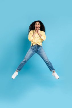 Excited Middle Eastern Lady Jumping Posing In Mid Air Holding Hands Near Face Expressing Excitement Over Blue Studio Background. Wow Offer Advertisement. Vertical, Full Length Shot