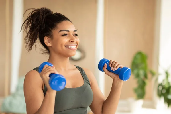 Fitness concept. Portrait of happy african american lady exercising with two dumbbells and smiling, enjoying domestic workout, free copy space.