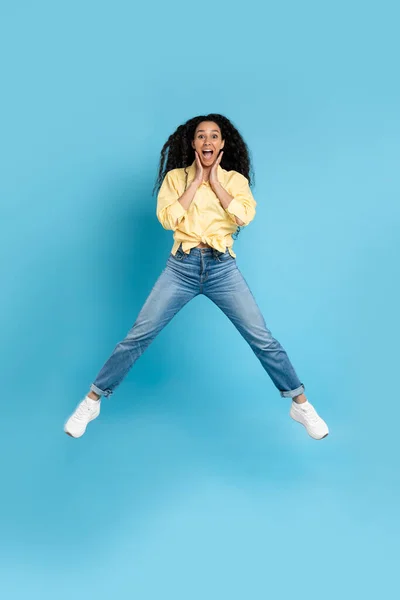 Excited Middle Eastern Lady Jumping Posing In Mid Air Holding Hands Near Face Expressing Excitement Over Blue Studio Background. Wow Offer Advertisement. Vertical, Full Length Shot