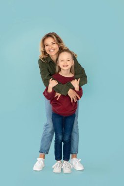 Vertical Shot Of Cheerful Mommy Hugging Her Kid Daughter Posing Standing Over Blue Background In Studio. Mother And Child Expressing Positive Emotions Advertising Offer