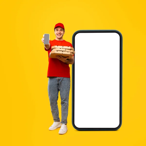 Pizza Delivery Guy Holding Boxes Showing Smartphone Standing Near Big Cellphone With Empty Screen Advertising Food Delivery App Over Yellow Background. Studio Shot, Square