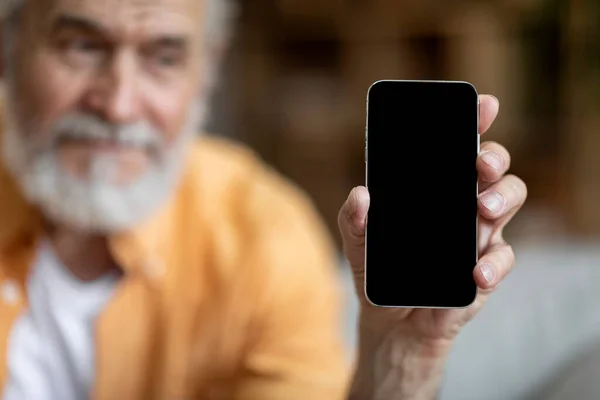 Cell phone with black empty screen in mature man hand, mockup. Positive bearded and long-haired elderly man sitting on sofa in living room, using mobile phone, showing nice offer, copy space, closeup