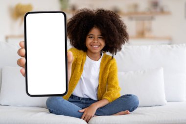 Adorable black little girl with cute bushy hair sitting on sofa at home, showing modern cell phone with white empty screen and smiling, recommending mobile app, mockup for advertisement, copy space