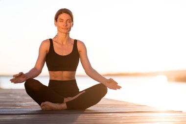 Wellness Concept. Smiling Middle Aged Female Meditating Outdoors On Wooden Pier, Relaxed Calm Woman Sitting On Yoga Mat In Lotus Position With Closed Eyes, Practicing Meditation On Sunset, Copy Space