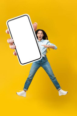 Shocked Arabic Lady Showing Big Phone With Empty Screen Jumping And Advertising New Mobile Application Standing Over Yellow Studio Background, Looking At Camera. Vertical