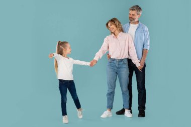Kid Daughter Pointing Finger At Camera Holding Hands With Parents Posing Together Over Blue Studio Background. Happy Family Choosing You, Advertising Great Offer. Full Length