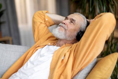 Relaxed grey-haired bearded elderly man in casual outwear sitting on couch, leaning back, enjoying newest wireless headset, listening to music at home, copy space, closeup shot, side view