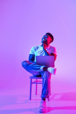 Thoughtful young black guy sitting on chair with modern laptop, looking at empty space above his head in neon light, full length. African American man having online job or business meeting