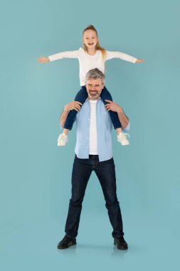 Vertical Shot Of Daddy Holding Little Daughter On Shoulders Posing And Having Fun Over Blue Studio Background, Smiling To Camera. Family And Parenthood Concept. Full Length