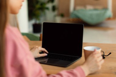Schoolgirl using laptop with blank screen, learning and doing homework, typing on keyboard while sitting at desk at home. E-learning, educational website advertisement