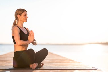 Zen Concept. Beautiful Middle Aged Woman Meditating On Pier Outdoors, Relaxed Female In Activewear Sitting In Lotus Position With Clasped Hands, Practicing Yoga Outside Near River Or Lake, Copy Space