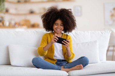 Happy smiling african american little girl with bushy hair sitting on couch with smartphone, cheerful kid playing mobile games, using nice application, home interior, copy space