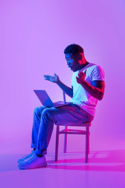 Deadline, online work stress. Portrait of emotional young black man sitting on chair with laptop computer, feeling shocked, making mistake in business project in neon light, full length. Copy space