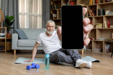 Cheerful handsome sporty senior man sitting on yoga mat, showing modern cell phone with black empty screen, recommending nice fitness mobile application, home interior, copy space, mockup