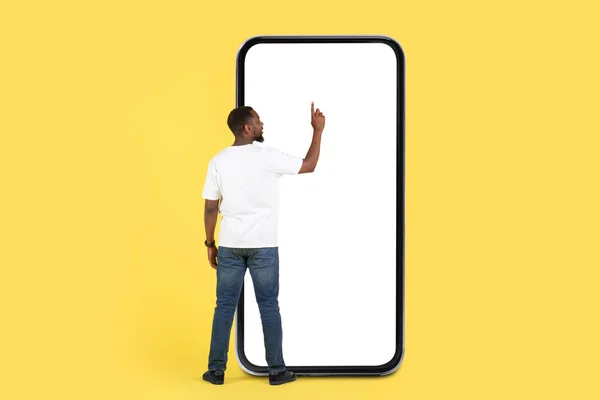 Back View Of Black Man Using Huge Phone With Empty Screen Standing On Yellow Studio Background. Guy Using Mobile Application And Browsing Internet On Big Smartphone. Mockup