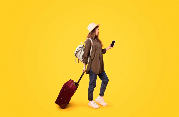 Tourist Lady With Suitcase And Backpack Using Phone Booking Tickets Online In Travel Application Online Standing Over Yellow Studio Background, Wearing Summer Hat. Technology And Tourism