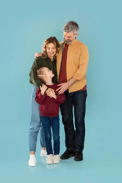 Full Length Shot Of Loving Family. Middle Aged Parents Embracing Daughter Smiling Standing Together In Studio On Blue Background. Parenthood And Childhood Concept. Vertical