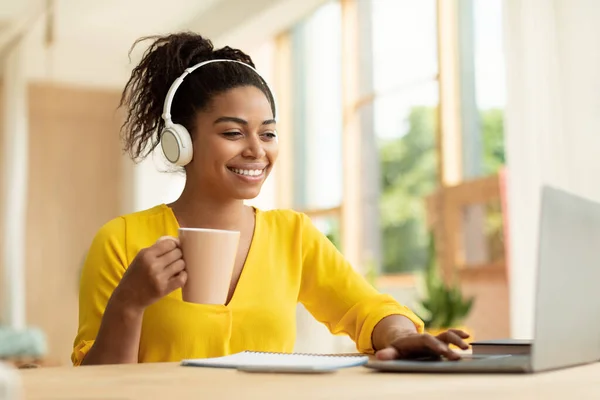 Happy african american lady in headphones working on laptop, holding cup and drinking coffee, sitting at desk at home. Smiling woman watching video, movie or webinar, taking break