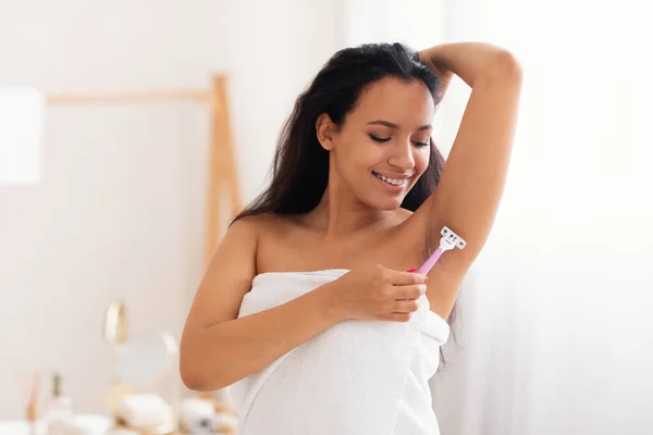 Cheerful Young Lady Shaving Armpit Hair Using Safety Razor In Bathroom At Home. Woman Making Depilation Standing Wrapped In Towel. Hair Removal Cosmetics Concept