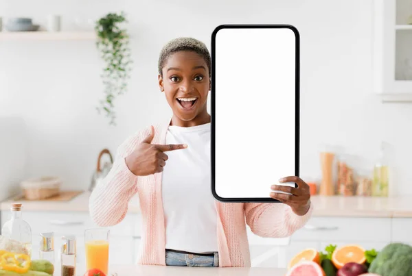 Excited Black Lady Holding And Pointing At Blank Big Smartphone In Kitchen, Emotional Young African American Woman Recommending New Mobile Application Or Website, Creative Collage, Mockup