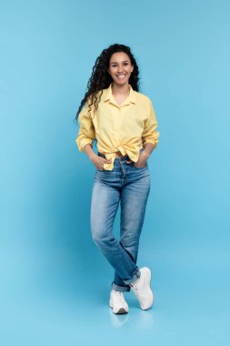 Full length portrait of positive young woman in trendy casual outfit standing and looking at camera over blue studio background. Happy millennial lady in shirt, jeans and sneakers smiling and posing