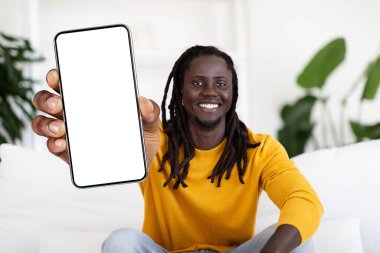 Cheerful Black Guy With Dreadlocks Showing Big Blank Smartphone With White Screen At Camera, Happy African American Man Recommending Mobile App Or Website, Sitting On Couch At Home, Collage, Mockup