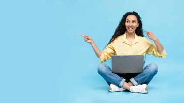 Smiling Middle Eastern Lady Using Laptop And Pointing Fingers Aside At Empty Space For Text Sitting On Blue Studio Background. Look There, Offer Advertisement Concept. Panorama
