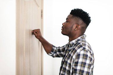 Black Millennial Man Knocking At Closed Door Of Apartment Indoors. Man Standing Near Entry Of Flat Posing In Doorway. Real Estate Business And Offers Concept. Side View Shot
