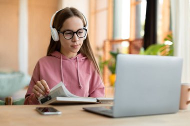 Focused female teenager studying, reading notebook and looking at laptop screen, learning remotely from home. Interested teen girl getting ready for online test or exam