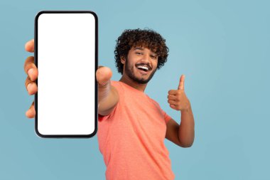 Modern smartphone with white blank screen in positive curly, bearded indian guy hand, positive eastern man showing nice mobile application and thumb up, blue background, mockup, copy space