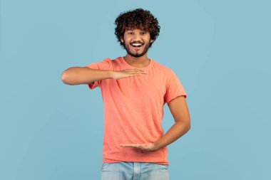 Cheerful handsome curly young indian guy in pink t-shirt showing or holding something invisible in his hands and smiling at camera over blue studio background, copy space