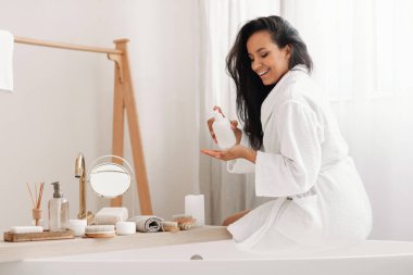 Joyful Attractive Female Caring For Body Holding Moisturizer Balm Bottle Sitting On Bathtub Wearing Bathrobe In Modern Bathroom At Home. Beauty Care, Skincare Cosmetics Concept