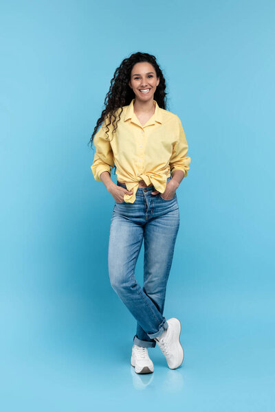 Full length portrait of positive young woman in trendy casual outfit standing and looking at camera over blue studio background. Happy millennial lady in shirt, jeans and sneakers smiling and posing