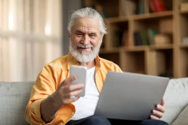 Happy bearded old man in stylish casual outfit sitting on sofa in living room, using brand new smartphone and laptop, having fun at home, copy space. Modern technologies and seniors concept