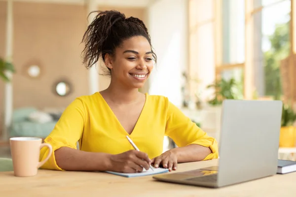 Smiling african american lady working on laptop taking notes in notebook, sitting at desk at home. Happy black female watching webinar on computer or writing check list, copy space