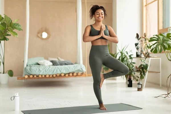 Calm black lady in sportswear exercising at home, standing in tree pose, meditating on fitness mat in bedroom interior, copy space. Healthy, sporty lifestyle, yoga practicing at home