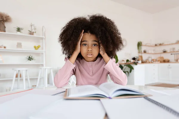Worried african american school girl with bushy hair sitting at desk full of books and notepads, stressed kid pupil doing homework alone, touching her head, copy space. Difficulties with education