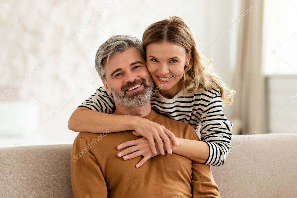 Happy Middle Aged Wife Hugging Husband Standing Behind His Back Sitting On Couch At Home. Loving Couple Embracing Posing Indoors. Marriage And Romantic Love Concept