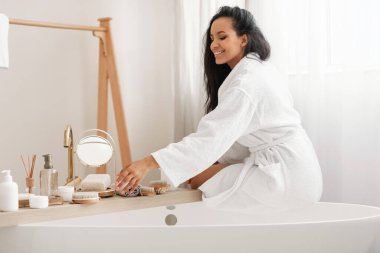 Beauty Care. Happy Lady Caring For Herself Using Cosmetics Products Sitting On Bathtub In Modern Bathroom At Home, Wearing Bathrobe. Wellness And Spa, Bodycare Concept
