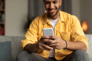 Happy young black male using smartphone, browsing internet, posting in social media at home, selective focus. Cheery millennial African American guy messaging with friend on mobile device